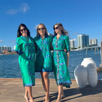 Three women in matching green outfits standing on a dock with water and city skyline in the background.