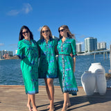 Three women in matching green outfits standing on a dock with water and city skyline in the background.