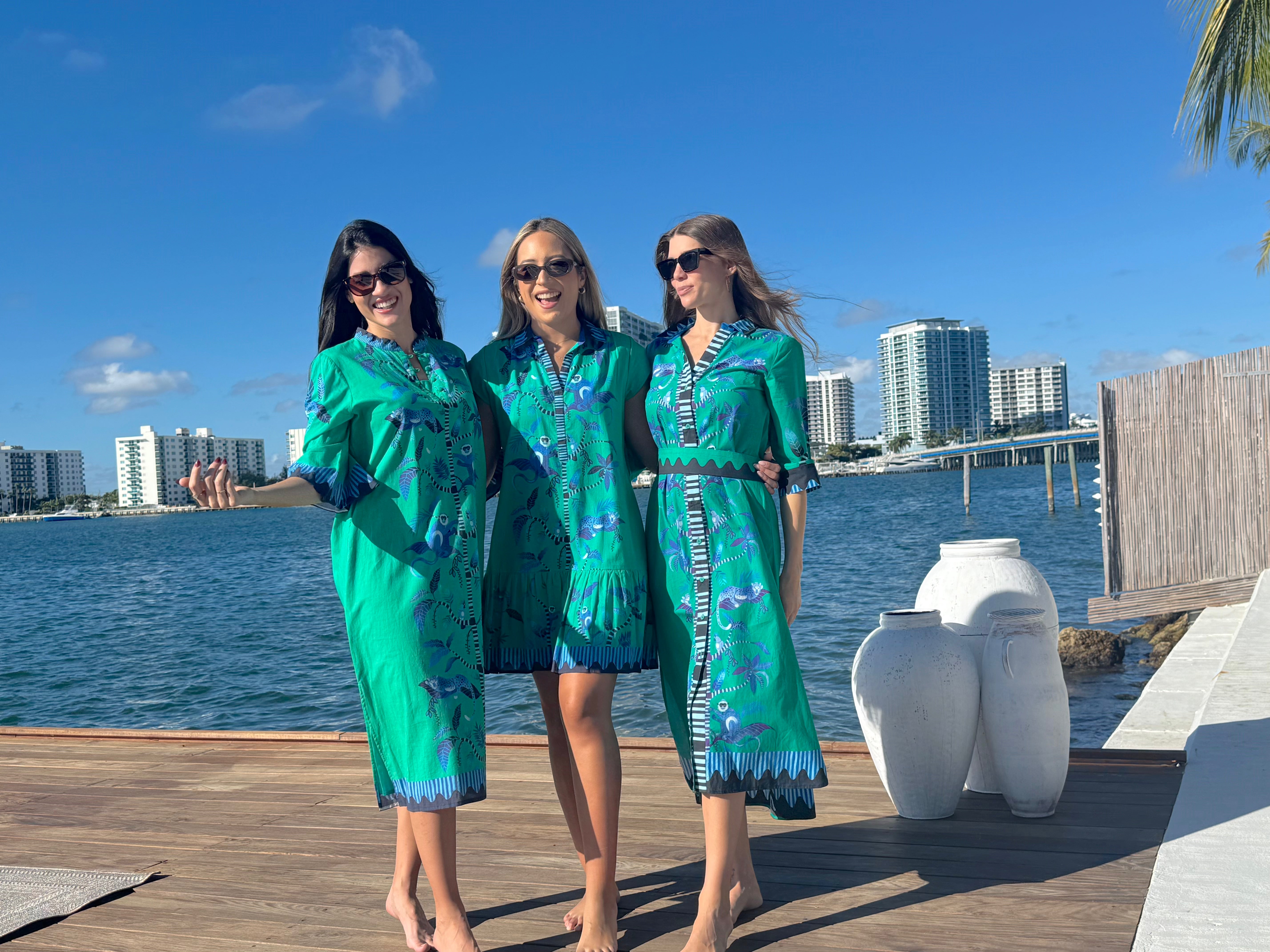 Three women in matching green outfits standing on a dock with water and city skyline in the background.