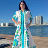 Woman in a white dress with blue and green patterns standing on a dock by water with buildings in the background.