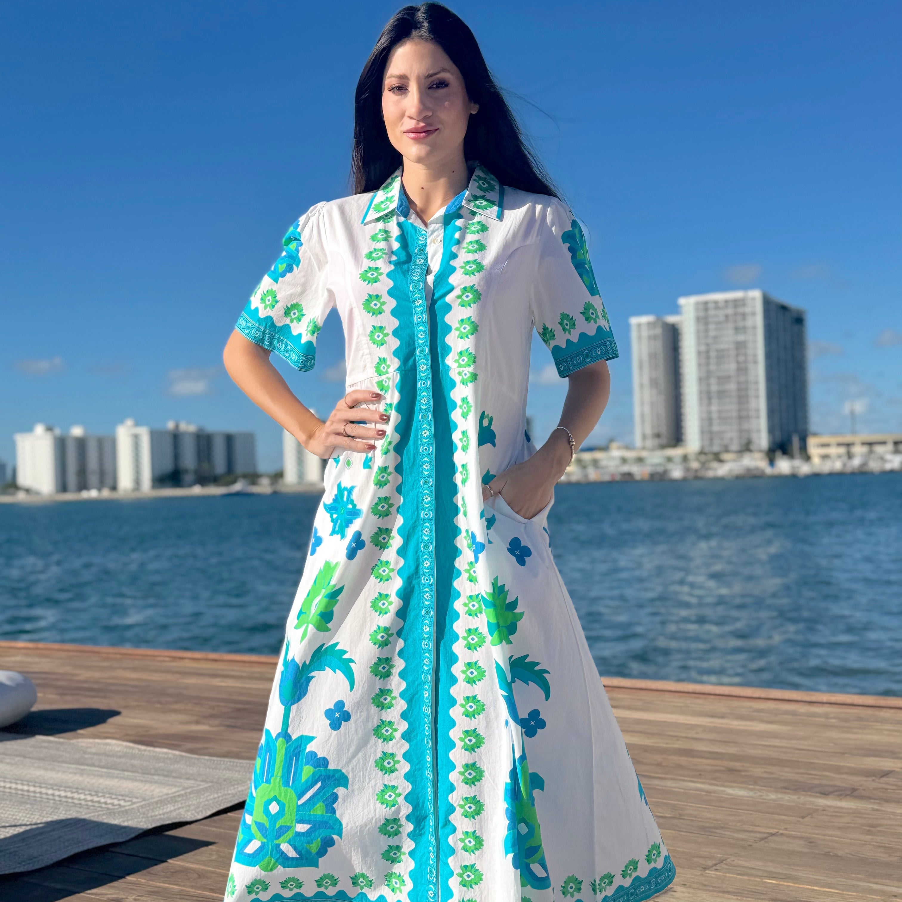 Woman in a white dress with blue and green patterns standing on a dock by water with buildings in the background.