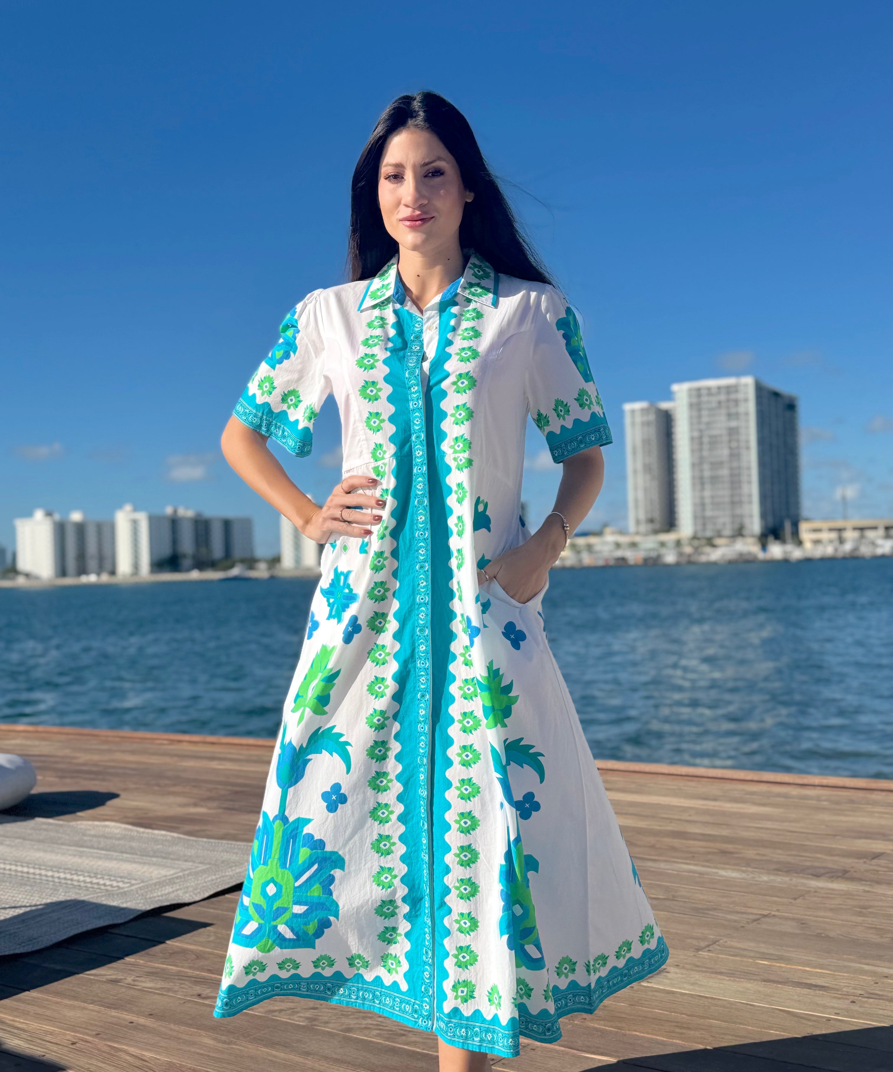 Woman in a white dress with blue and green patterns standing on a dock by water with buildings in the background.