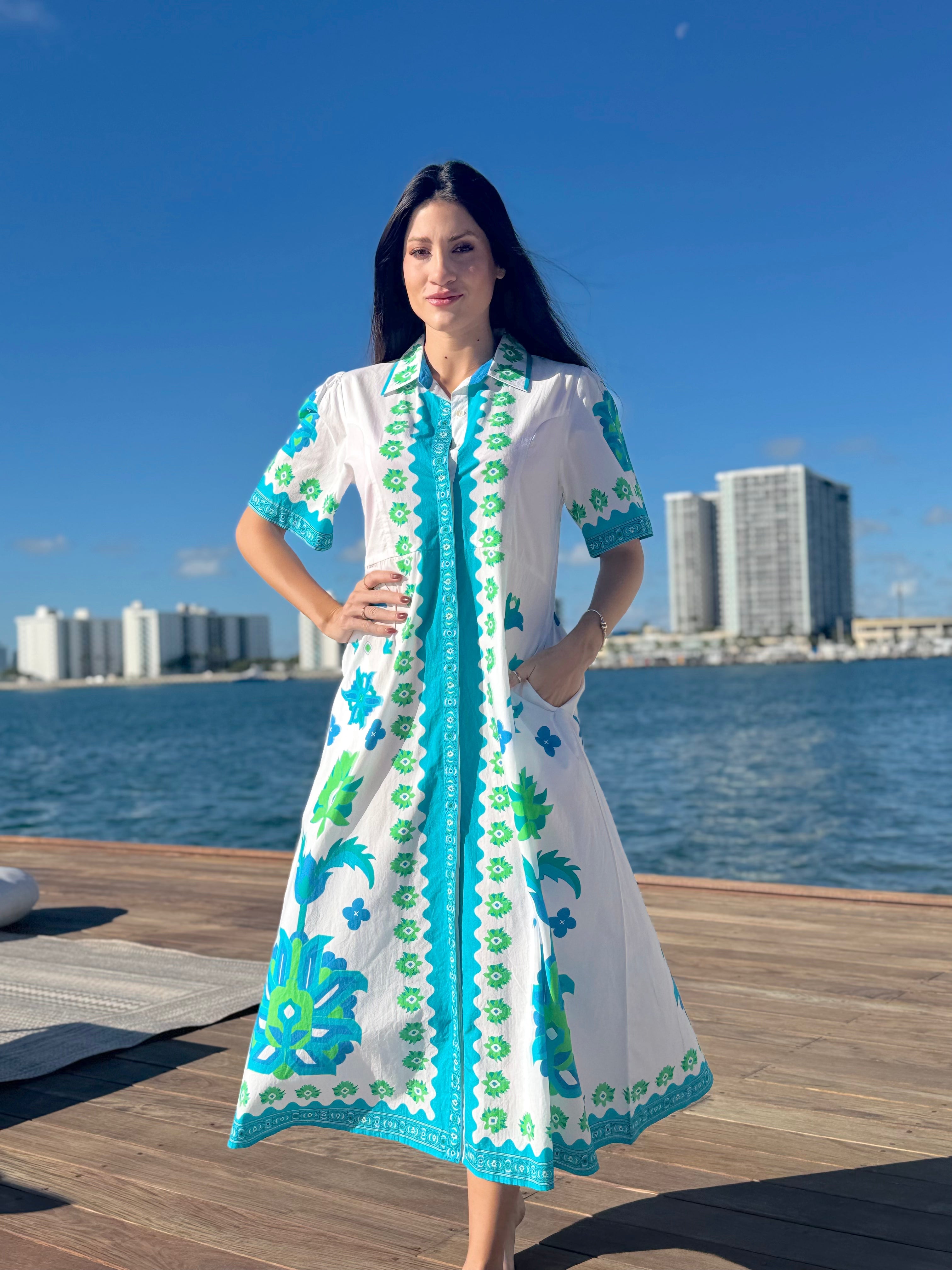 Woman in a white dress with blue and green patterns standing on a dock by water with buildings in the background.