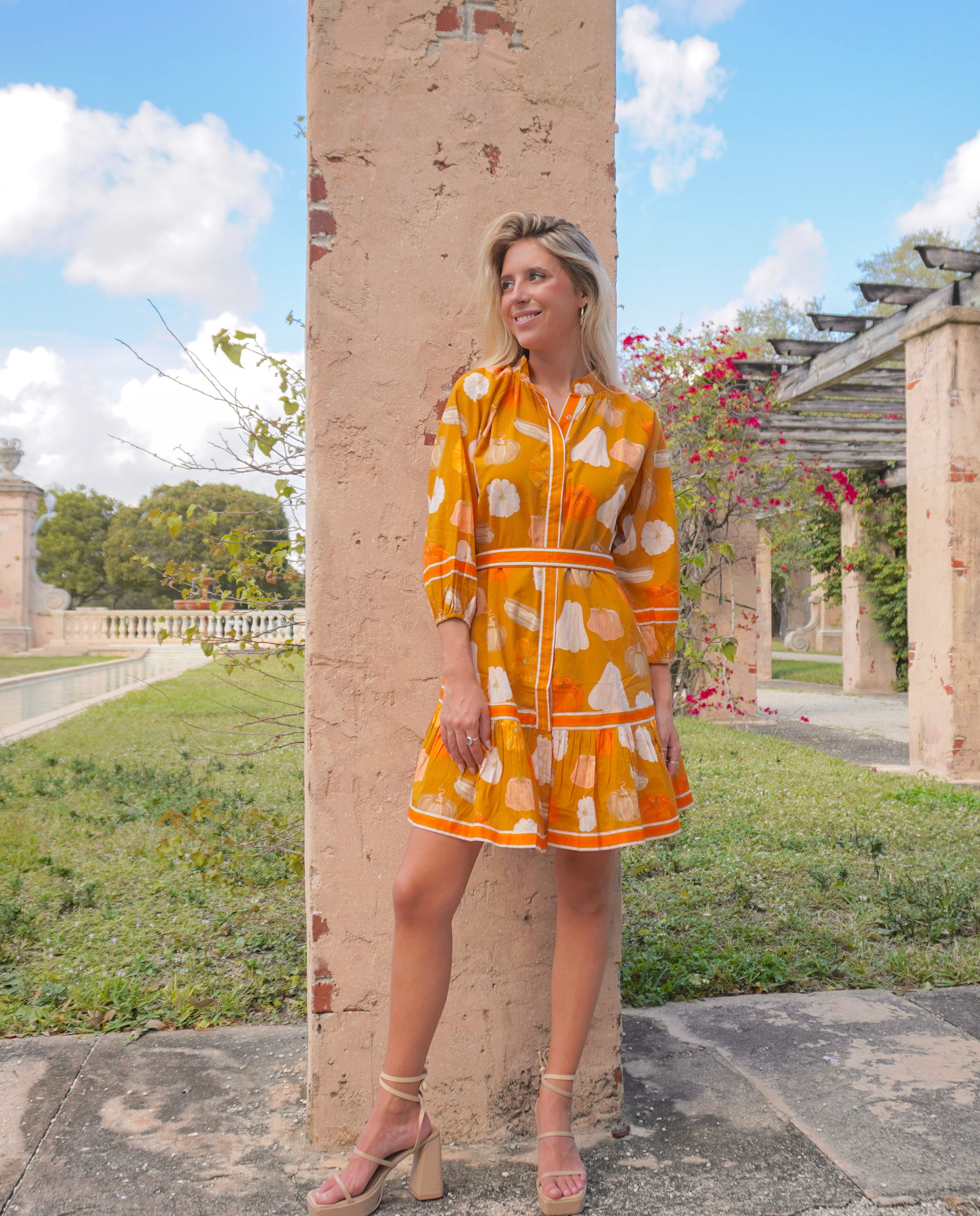 Woman in a yellow dress with a floral pattern standing outdoors near a stone column.